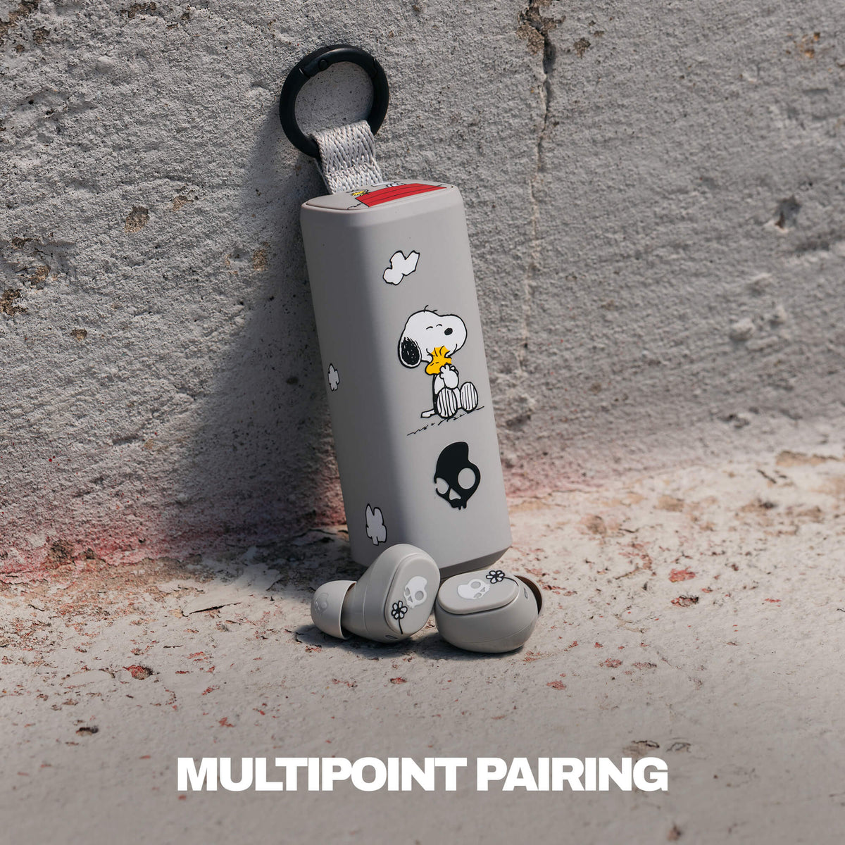 A pair of gray true wireless earbuds with a matching cylindrical charging case featuring woodstock illustrations, leaning against a textured concrete wall. The text 'MULTIPOINT PAIRING' is displayed at the bottom.  | Skullcandy X Peanuts Woodstock