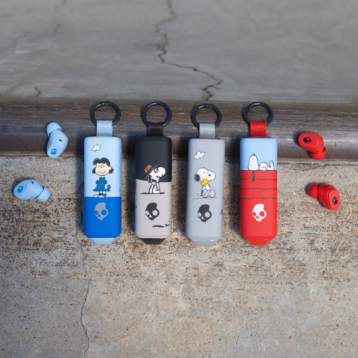 Four peanuts-themed dime 360 earbuds in blue, black, gray, and red are lined up on skate coping, with matching red and blue earbuds resting on either side on a concrete surface. | Skullcandy X Peanuts Woodstock
