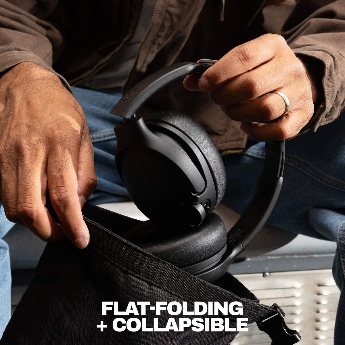 A close-up of a person holding a black over-ear headset over an open black bag, with the text 'FLAT-FOLDING + COLLAPSIBLE' at the bottom. The person is wearing a brown jacket and blue jeans. | Cement